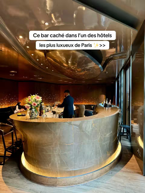 An interior shot of a luxurious bar. The focal point is a curved bar with a marble-like finish and a warm, inviting glow from a light that runs along its base. Behind the bar, a person in a dark suit is visible. Elegant decor, featuring gold and the lighting, creates a sophisticated ambiance. On the left side of the frame, there are bar stools and a glimpse of a seated figure, with a large floral arrangement adding a touch of color. The ceiling is reflective, with lights that create an interesting visual