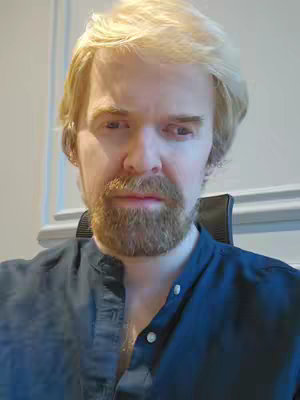 A close-up shot of a man with light blonde hair and a neatly trimmed, reddish-brown beard. He is wearing a dark blue collared shirt. The background is a neutral, possibly a wall in a room.