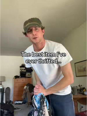 A man in a camo cap and white t-shirt stands in a room, holding a bundle of clothing and gesturing toward the camera. Text overlay indicates the content is about a thrifted item.