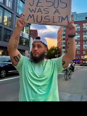 A man stands on a street holding a sign with text on it. He appears to be outdoors, near buildings and other cars. The sign says "JESUS WAS A MUSLIM". There are other people and cars that pass behind him.