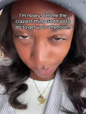 The cover image focuses on a Black woman from a close-up, head and shoulder view. She's wearing a gray hoodie over her head, with her hair falling in loose waves around her face. The woman has a serious expression, slight squint, and slightly arched lips. In the foreground, text overlayed with white font reads: 'I'm nosey. Tell me the craziest thing God had to do to get your attention..'. Her attire includes a white shirt underneath, with a gold heart pendant and a faint necklace. The background is a soft blurry texture.