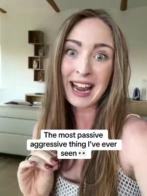 A woman with long blonde hair is shown up close with a surprised expression, wide eyes, and open mouth. She is wearing a white top and stands in a well-lit room with neutral-colored walls and some decor in the background.