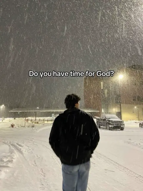 A man in a black jacket and jeans walks away from the camera in a snowy environment. Heavy snowfall is visible, with a building and streetlights in the background. The image has a cold, slightly desaturated color palette.