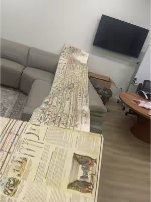 The image shows a room interior with a neutral color palette. A gray sectional sofa, a large flat-screen television mounted on the wall, and parts of a wooden table are visible. A long, rolled-up document with ancient hieroglyphics is draped over the sofa.