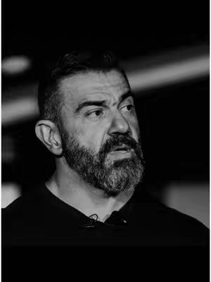 A black and white portrait of a middle-aged man with a beard, looking slightly to the side. The background is dark, and the lighting is focused on the man's face.