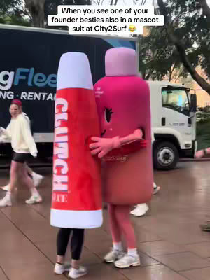 Two people in mascot costumes hug each other. The mascot on the left is shaped like a tube of lipstick with the brand 'CLUTCH' written on it. The mascot on the right is a pink capsule-shaped character with a smiling face. In the background, there is a white truck and a person walking with a white hoodie.