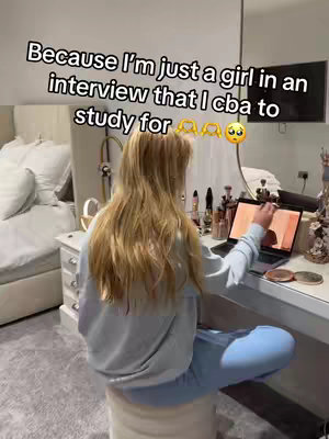 A woman with long blonde hair sits in front of a vanity table in a well-lit room. She is facing away from the camera, looking at a laptop screen. The room is decorated in neutral tones with a large mirror and a bed visible in the background.