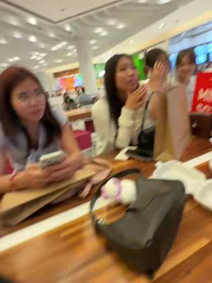 The image shows a group of young women at a brightly lit cafe or shopping center. The women are seated at a wooden table, the foreground is dominated by a black purse. The women are looking in different directions, some of them are holding phones. The overall impression is that of a casual gathering, possibly a social event.