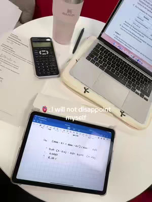 A top-down view of a study setup on a white table. An open laptop displays text on the screen. A calculator is on the table, next to a pen and a large pink tumbler. A tablet showing notes and equations is towards the bottom. The image is lit with bright, natural light.