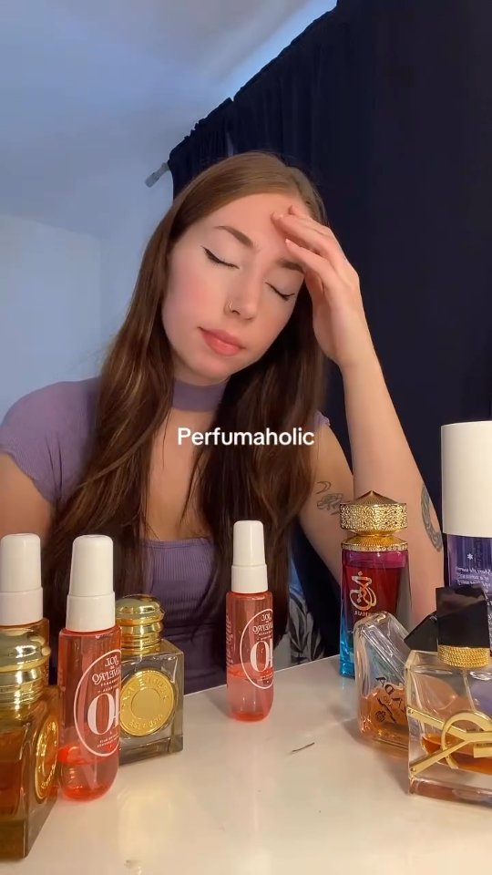 A young woman is seated at a table, with a table full of perfumes in the foreground. She is looking downward with her eyes closed and her hand resting on her forehead in a gesture of contemplation. She is wearing a purple top. The overall setting seems to be her room. The background includes a curtain.