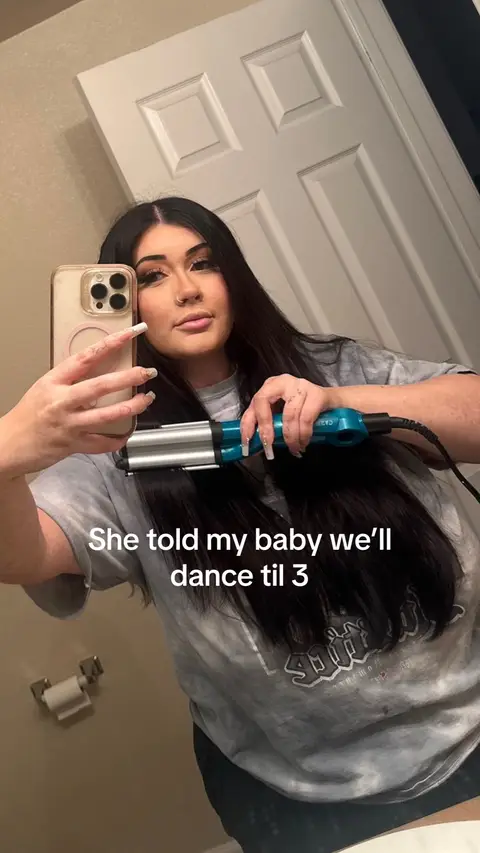 A woman is holding a hair crimper in front of a mirror, framed in a selfie. She appears to be in a bathroom as other items are visible in the background. The text overlay says "She told my baby we'll dance til 3".