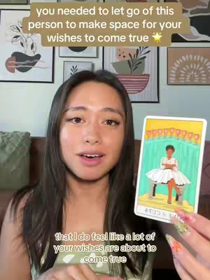 A young woman with long dark hair is centered in the frame, holding a tarot card and speaking. Behind her are framed art pieces on the wall. A light-colored text overlay provides additional context at the top.