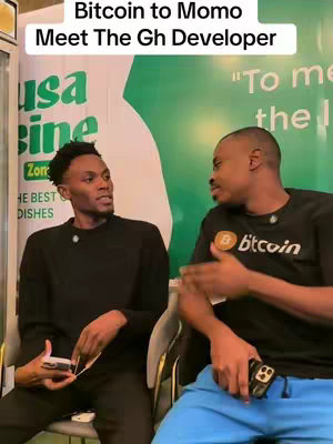 Two young Black men converse indoors. The man on the left, in a black shirt, holds a phone and looks towards the right while speaking. The man on the right, wearing a black t-shirt with a Bitcoin logo, gestures with his hands. Behind them is a green wall with some logos.