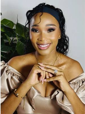 A smiling woman in a golden dress posing with her hands together. She has a black headband, and her hair is styled in curly waves. Her makeup is glamourous.