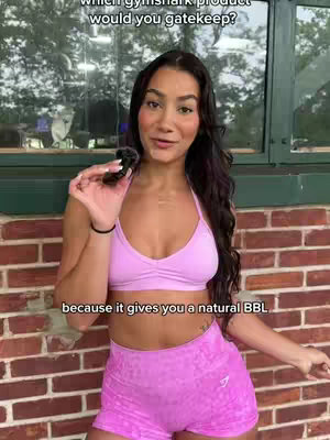 A woman with long dark hair is holding a small black object with a pink top and shorts in front of a red brick wall. The image presents a medium shot with the woman centered, the background including a window and foliage, overlaid with text at the top and bottom.