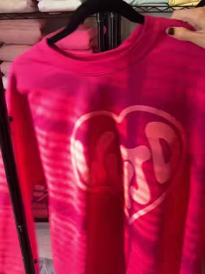 The cover image shows a brightly colored, pink crewneck sweatshirt hanging on a rack. The sweatshirt has a large heart graphic in the center, and inside the heart is the visible text. The background is a bit blurred, with shelves and other clothing items suggesting a retail setting.