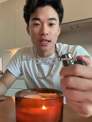 A man in a white t-shirt is holding a lighter next to a burning candle in a glass jar. In the foreground is the candle with a lit wick and a pool of melted wax at the top. The man is looking at the camera with an expression of slight amusement. Below the man's face is overlaid text. The background is out of focus but appears to be a kitchen setting.