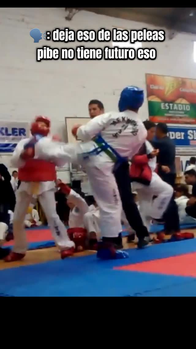 The image shows a martial arts match in progress. Two figures in Taekwondo uniforms are engaged in combat. The foreground features a blue mat, and the background includes spectators and event signage.