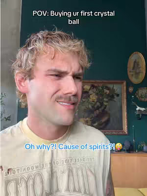 The image captures a man with light skin and light-colored, wavy hair. He has a mustache and a slightly confused or questioning facial expression. Text overlays include “POV: Buying ur first crystal ball” above his head and "Oh why?! Cause of spirits?!" below, along with a smiling emoji. The background features a dark teal wall, a framed picture, and decorative items.