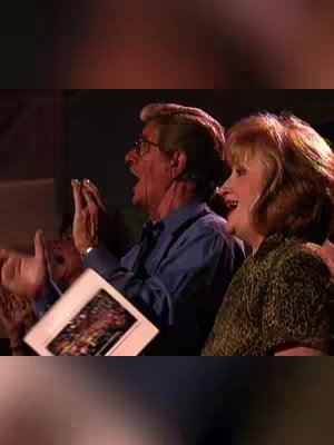 A snapshot from a concert, likely from the Gaither Vocal Band, shows an older man and woman in the audience with joyful expressions. The man is clapping with both hands raised, while the woman, with short blonde hair, has an open mouth as if singing along. They are closely framed, suggesting an intimate view of their experience, with a hint of lights in the background.