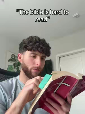 A man is shown holding a Bible with a bookmark. Text overlay reads, 'The bible is hard to read'. The man is fair-skinned with dark curly hair, a beard, and wearing a grey shirt. The setting is domestic, with plain walls and a door visible behind him.