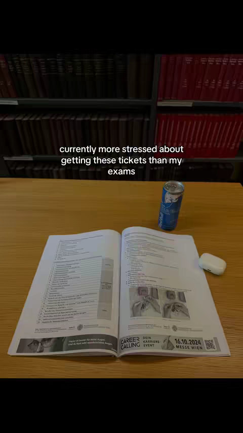 An open textbook with text and images is centrally placed on a wooden desk. A can of blue liquid is next to the book. In the background a bookshelf lined with books. Text overlayed towards the top: 'currently more stressed about getting these tickets than my exams.'