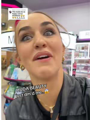 A woman with wide eyes and an open mouth appears surprised and excited, centered in the frame. She's in what looks like a makeup store, with shelves and products in the background. Text overlays: 'HUDA BEAUTY just dm'd me..'