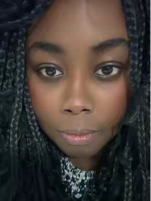 A close-up shot of a person's face, likely a selfie. The subject has dark skin, dark eyes, and is wearing their hair in multiple braids. The background is out of focus.