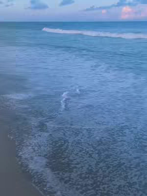 The image shows a serene beach scene. The foreground features a sandy shore, while the midground displays the ocean with gentle waves lapping against the beach. The water transitions from a darker blue to a lighter shade as it forms waves closer to the shore. The sky is a light blue with soft, fluffy clouds tinged with pink, suggesting either sunrise or sunset. The overall color palette is cool and soothing, creating a calming atmosphere.