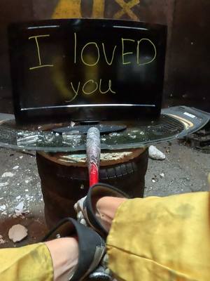 A person holding a baseball bat, standing next to a broken television set that is resting on what appears to be a tree trunk, in an outdoor dark environment. The televisions screen has the handwritten phrase, "I LOVED you". The person is wearing yellow clothing and black shoes.