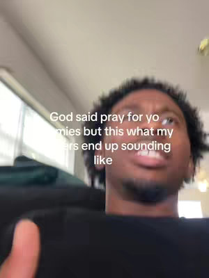 The image is a selfie-style video still featuring a young, Black man filmed from a low angle. The man is in focus, with his head tilted slightly upwards and to the right. He has short, dark, curly hair and is wearing a black shirt. the shot appears to be taken indoors, with a light-colored wall or ceiling visible in the background and to the left.
