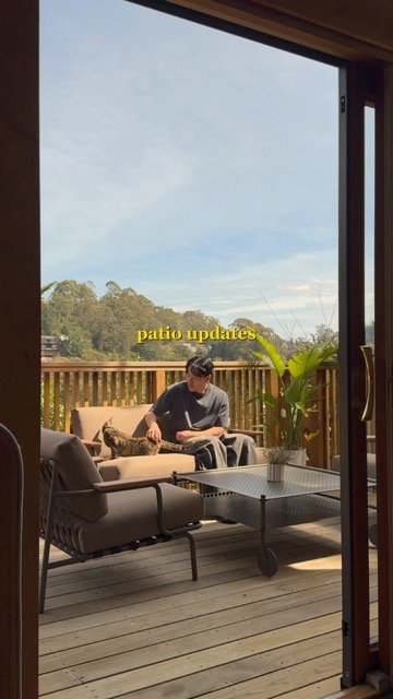 A person is sitting on an outdoor sofa, petting a cat. The scene is framed by a sliding glass door. In the background is a wooden deck with trees and a blue sky.