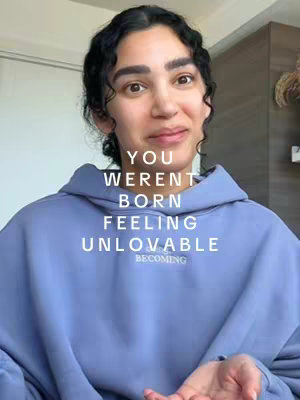 A medium shot features a woman in her late twenties or early thirties with light skin, dark curly hair, and brown eyes, smiling warmly at the camera. She's wearing a pale blue hoodie. Above her and in the lower-mid section of the frame is white text that reads, "YOU WEREN'T BORN FEELING UNLOVABLE". The background is somewhat blurred, suggesting an indoor setting with a hint of a light-colored door or wall.