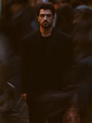 A man stands center in an image, facing the viewer. He is surrounded by blurred figures in the background that suggests a crowd. He has short, dark hair and a beard and is wearing a dark outfit. The lighting is dim overall, with the background being out of focus.