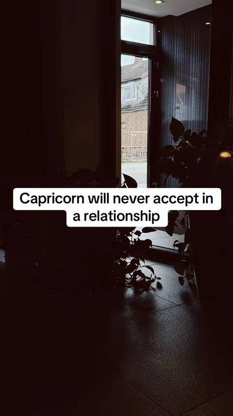 An interior shot with a dark, moody ambiance. A window is on the right side, with visible panes providing a view of the outside. A text overlay reading 'Capricorn will never accept in a relationship' is positioned at the center of the frame on a white rectangle.