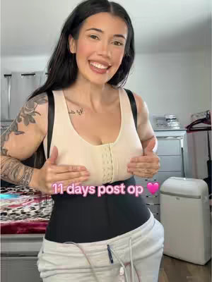 A woman is shown from the waist up, centered within the frame. She is wearing a light beige sports bra and a black compression garment around her midsection. She has dark hair and is smiling, with both hands placed on her chest, seemingly supporting her breasts. The background includes a glimpse of what appears to be a dresser and a white appliance. Overlaying the image is text that reads '11 days post op' in pink, and a pink heart.