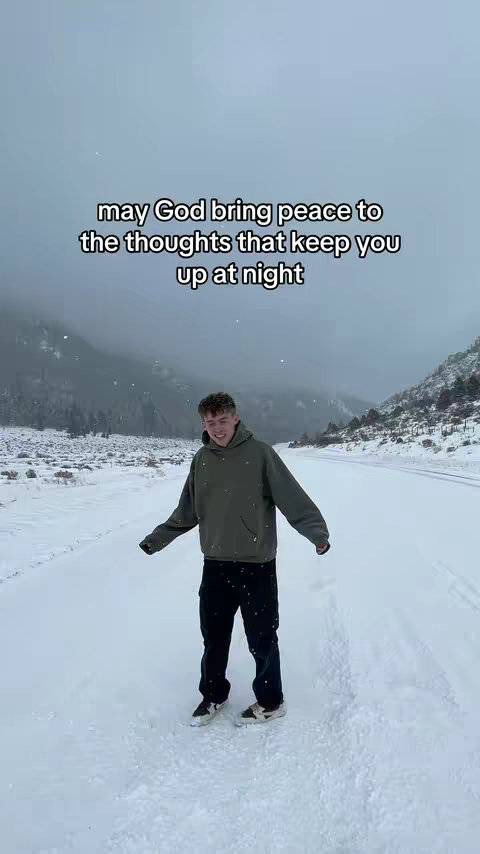 A young man stands on a snow-covered road, flanked by snowy landscapes and distant mountain ranges. The sky is overcast, creating a soft lighting.