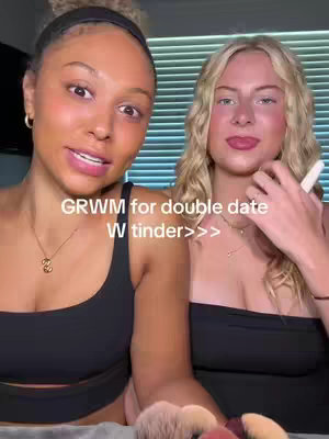 Two young women are in a selfie-shot, looking at the camera. The woman on the left has darker skin, dark hair pulled back and is wearing a black top and a necklace. The woman on the right has blonde hair , and is wearing a black top as well. The background shows horizontal blinds.