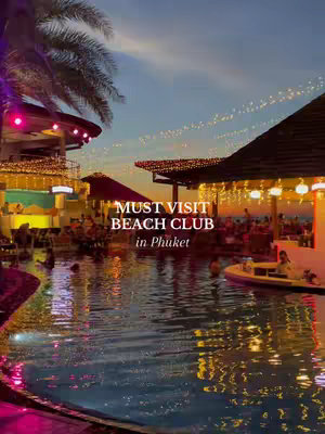 An eye-level shot captures the exterior of Kudo Beach Club in Phuket at dusk. The scene is dominated by a large pool of water in the foreground, reflecting the ambient lighting. The club is built around a central pool area, with outdoor seating and structures illuminated by soft, warm lights. The sky transitions from shades of blue to orange, and the overall composition is balanced with the water and structures creating a sense of symmetry.