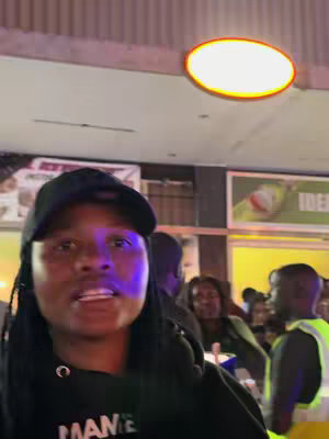 A selfie-style video shows a person, wearing a cap and hoodie, speaking in front of blurred background. Various people are behind them, and a sign in the back is also visible. The ceiling has a round illuminated light.