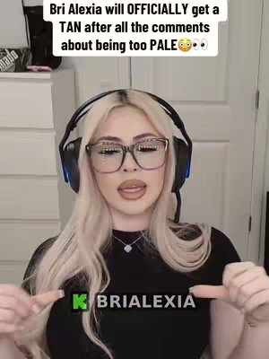 A woman with long blonde hair and glasses is in a medium shot, looking directly at the camera. She wears a headset and a black top with a logo, and her fingers point towards it. The background shows a neutral-toned wall and some objects that appear to be on a shelf.