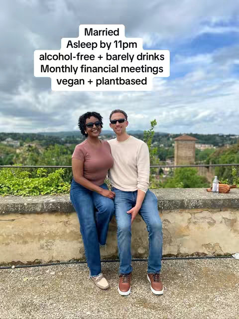 The cover image shows a couple sitting side-by-side on a stone wall, with a scenic background of a city and a cloudy sky. The female is on the left wearing a pink top and blue jeans. The male is on the right in a light-colored shirt and blue jeans. The text overlay in the upper center states: Married, Asleep by 11pm, alcohol-free + barely drinks, Monthly financial meetings, vegan + plantbased