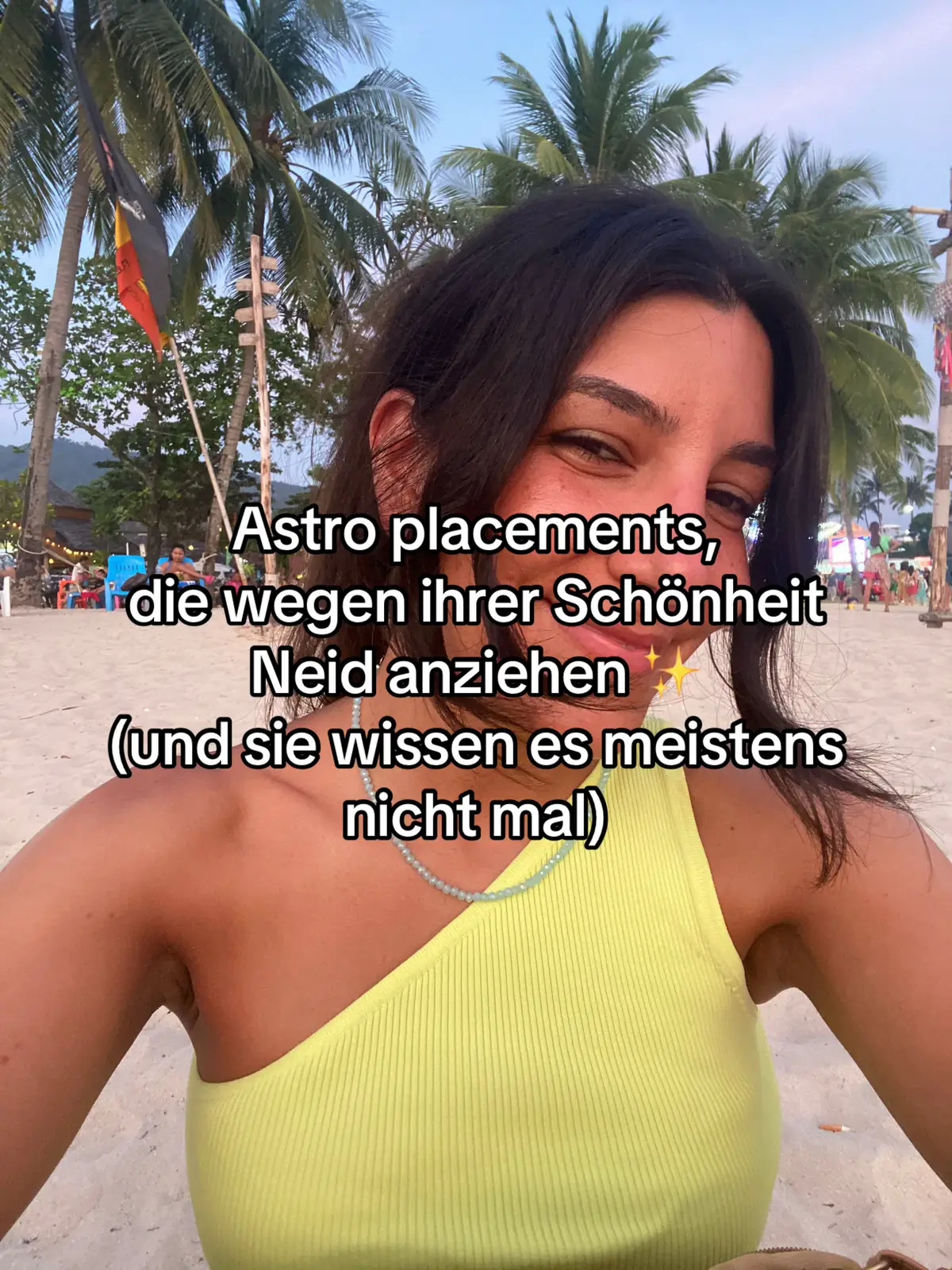 A selfie taken on a beach with the creator in the foreground. The creator is a person with dark hair and is wearing a yellow top. Palm trees are visible in the background, and text overlays the image.