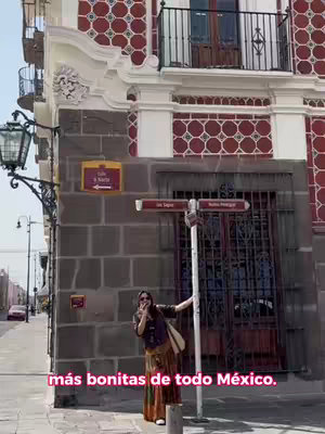 The cover image showcases a woman smiling while posing by a building in Puebla, Mexico. She is standing next to a signpost with street names, suggesting a place for tourist information. The background features a building with a red and white patterned facade and a wrought iron balcony. The woman is dressed in a casual bohemian style, wearing sunglasses. The overall scene conveys a sense of travel and exploration.