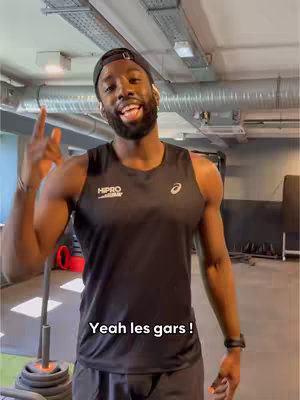 A man in a black tank top and a baseball cap is posing in a gym. The man is smiling with his mouth open, pointing with his index finger and extending his other hand with the peace sign gesture. The background shows gym equipment and a glimpse of the ceiling with air ducts.