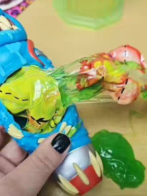 The image shows a person holding a blue and silver dissected toy alien figure with an open belly, revealing colorful internal contents. A hand with a black-polished fingernail is also visible, touching the toy. Green slime-like material spills from the toy onto a light brown surface.