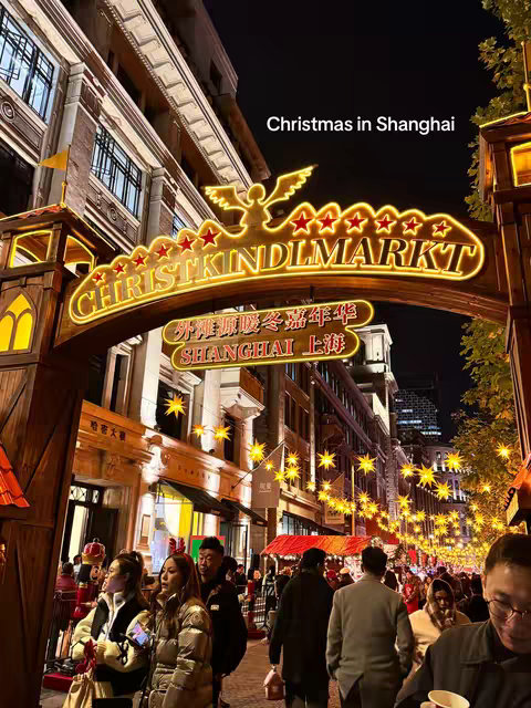 An eye-level night shot captures a vibrant Christmas market scene. The frame showcases a street or walkway adorned with twinkling star-shaped lights suspended overhead, leading the eye towards the brightly lit entrance, reading 'Christkindlmarkt'. Several individuals are present, navigating the market.