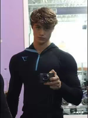 A young man wearing a black hoodie with light blue accents, holding a camera. The background is blurred, suggesting a gym setting with mirrors and equipment.