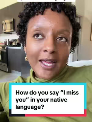 A woman with curly hair is shown from the chest up, looking slightly upward and to the side as if mid-conversation. She is wearing a green turtleneck sweater, and the background shows some of the interior of her home, including what looks like a kitchen area with a microwave and cabinets. A text overlay at the bottom contains the question, “How do you say 'I miss you' in your native language?”.