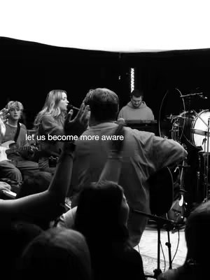 A black and white photo depicting a live music performance. A man in the foreground is seen from the back with his arms raised, facing the stage. On the stage, a band is performing, including a female vocalist at the microphone, a guitarist, a drummer, and a person at a keyboard. Additional people are in the background, suggesting an audience or more band members. The lighting is dim, with a spotlight effect on the performers.